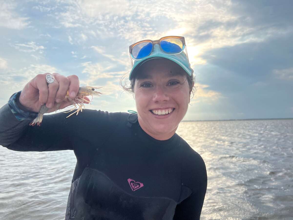 Dr. Lele Schlenker, a woman in a black wetsuit and blue ball cap, smiles while holding a shrimp in her hand. Blue sky and water appear behind her.