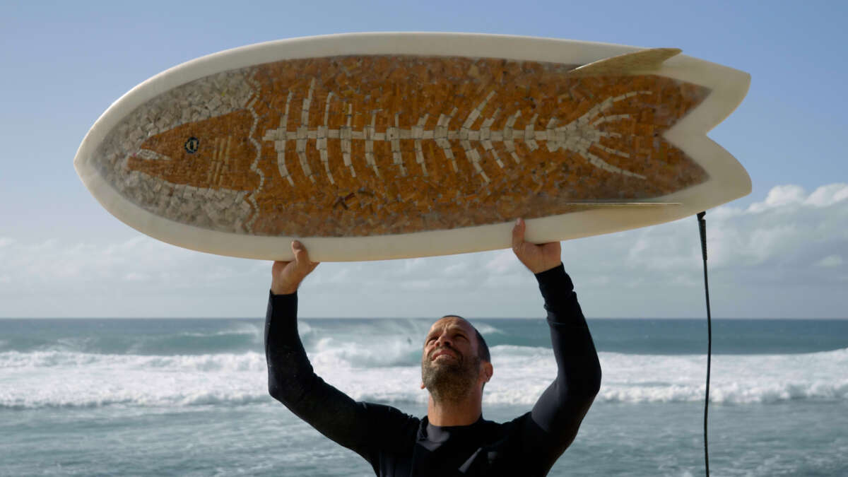 A man stands on the beach holding a cigarette surfboard horizontally above his head. Blue sky and the ocean are behind him.