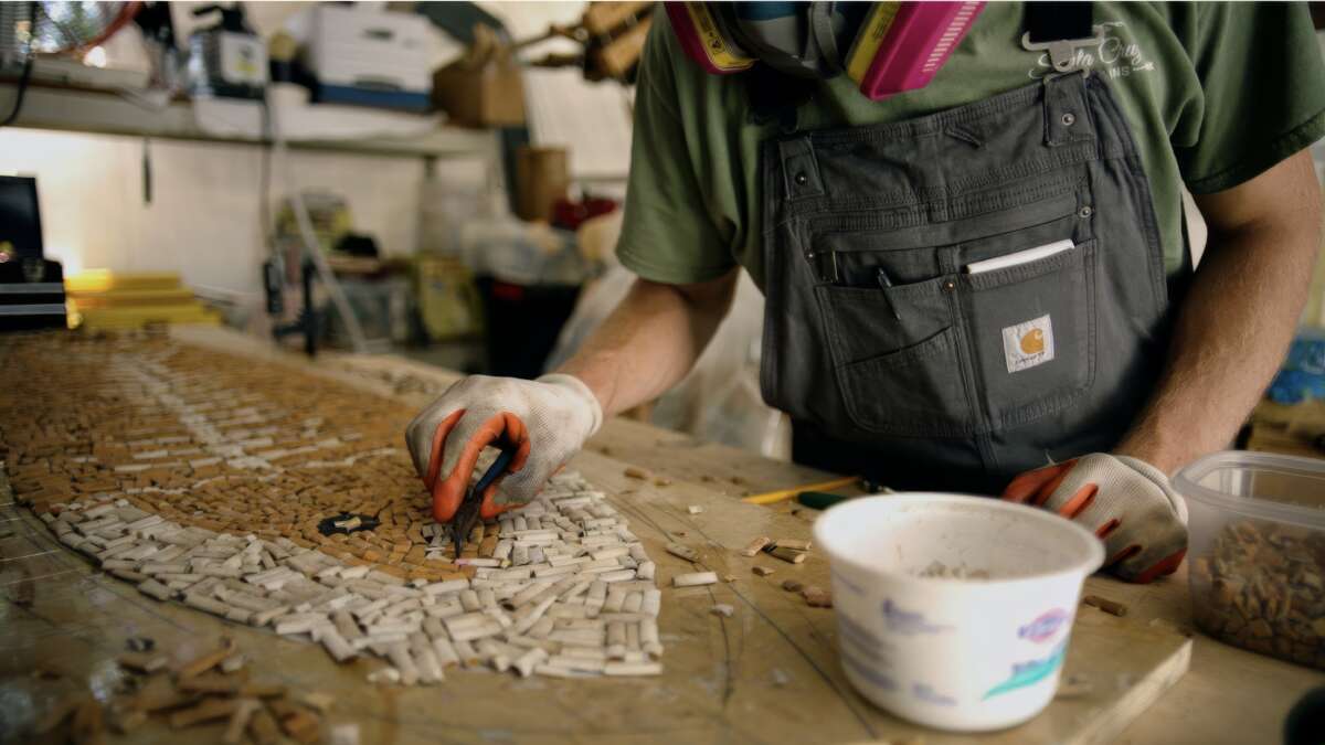 Some wearing grey overalls, a greentshirt, and orange and white gloves uses two fingers to place a cigarette in place during the construction of the surfboard.